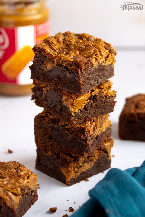 Biscoff brownies in a stack of 4 bars on a white work surface with a jar of Biscoff spread, a blue linen napkin and more Biscoff brownies in the background