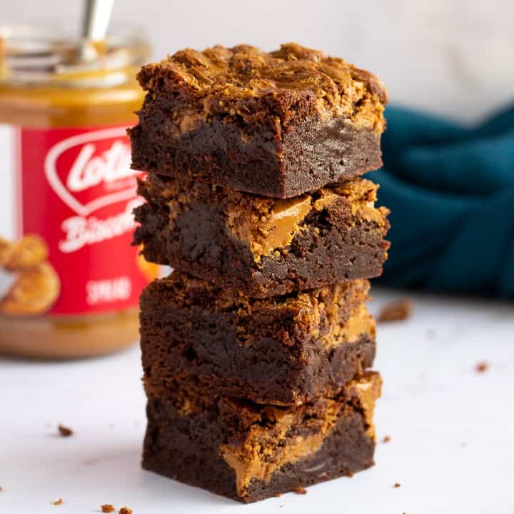 Biscoff brownies in a stack of 4 bars on a white work surface with a jar of Biscoff spread and a blue linen napkin in the background