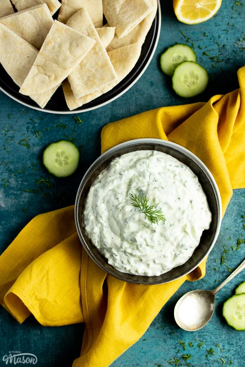 A bowl of tzatziki on a mustard yellow linen napkin with cucumber slices, half a lemon and pitta bread in the background