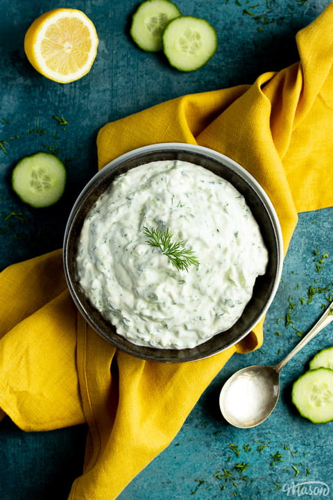 A bowl of tzatziki on a mustard yellow linen napkin with cucumber slices, half a lemon and a spoon in the background