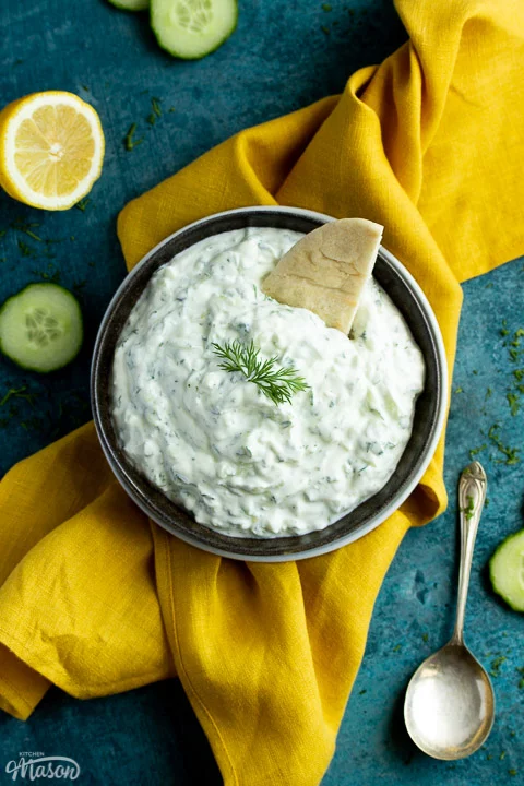 A bowl of tzatziki with pitta bread dipped in it on a mustard yellow linen napkin with cucumber slices, half a lemon and a spoon in the background