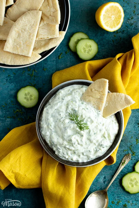 A bowl of tzatziki with pitta bread dipped in it on a mustard yellow linen napkin with cucumber slices, half a lemon and a spoon in the background