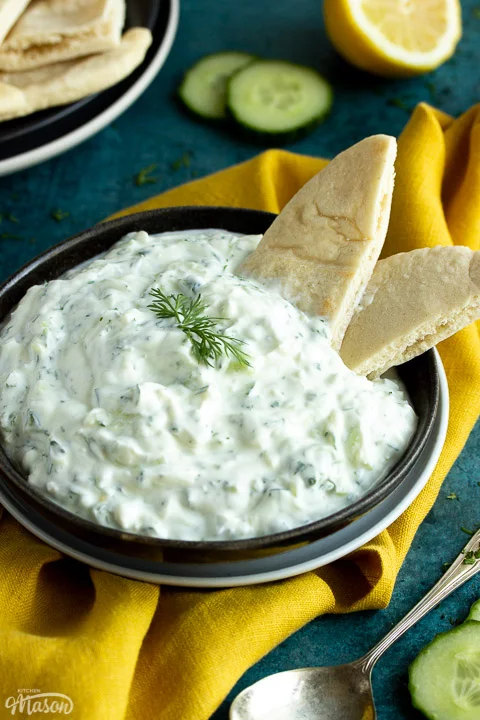 Close up of a bowl of tzatziki with pitta bread dipped in it on a mustard yellow linen napkin with cucumber slices, half a lemon and a spoon in the background