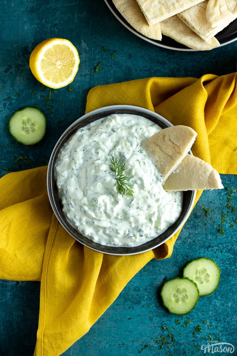 A bowl of tzatziki with pitta bread dipped in it on a mustard yellow linen napkin with cucumber slices, half a lemon and pitta bread in the background