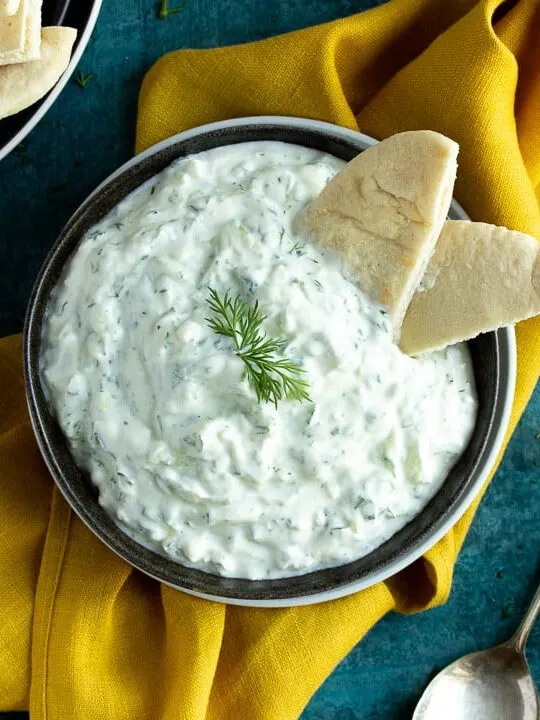 A bowl of tzatziki with pitta bread dipped in it on a mustard yellow linen napkin with cucumber slices, half a lemon, pitta bread and a spoon in the background