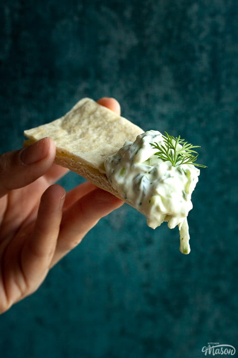 Close up of someone holding a pitta bread with a large dollop of tzatziki and a spring of fresh dill on the end
