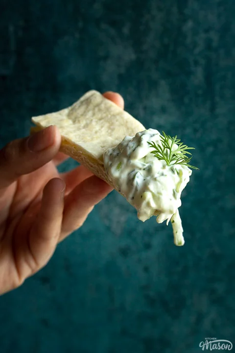 Close up of someone holding a pitta bread with a large dollop of tzatziki and a spring of fresh dill on the end