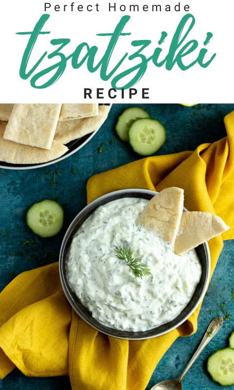 A bowl of tzatziki on a mustard yellow linen napkin with cucumber slices, pitta bread and a spoon in the background