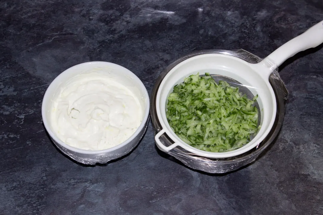 Grated cucumber in a sieve set over a metal bowl covered in cling film and the remaining tzatziki dip ingredients (minus the dill) mixed together in a small white bowl also covered in cling film.