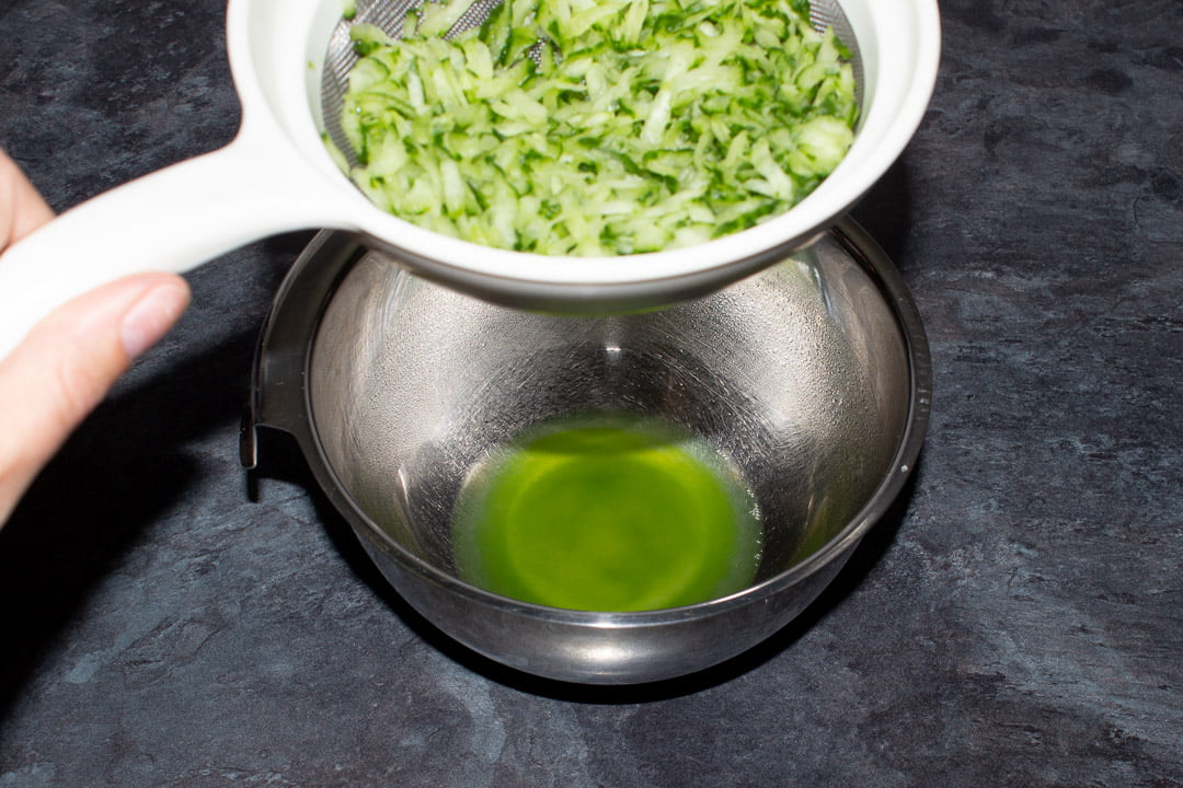 Grated cucumber in a sieve being held above a metal bowl containing cucumber juice.