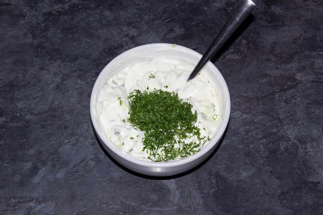 Freshly chopped dill being added to tzatziki in a small white bowl with a metal spoon
