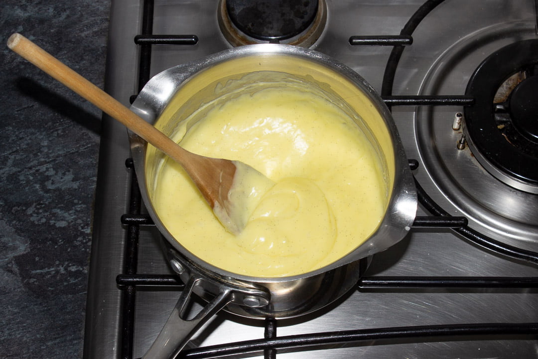 Thickened custard in a metal saucepan over a stove with a wooden spoon