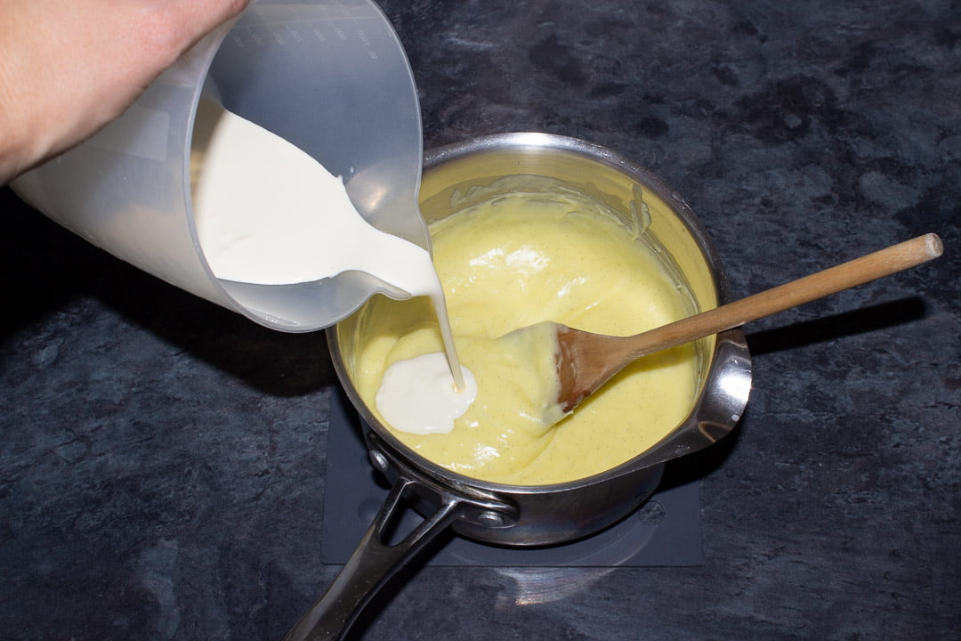 Cream being poured into a metal saucepan containing thickened custard and a wooden spoon