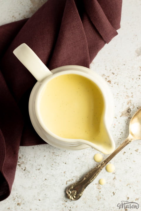 Homemade custard in a white jug on a burgundy linen napkin with drips running down the front and a silver spoon in the background
