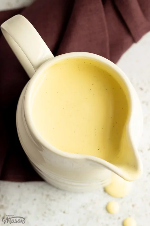Homemade custard in a white jug on a burgundy linen napkin with drips running down the front