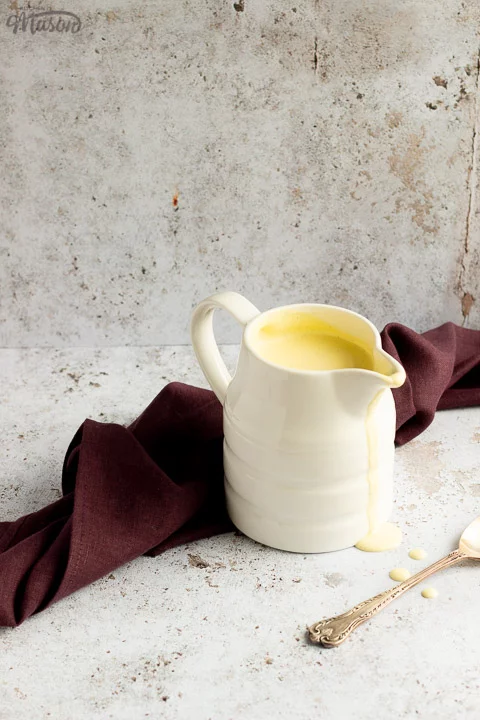 Homemade custard in a white jug on a burgundy linen napkin with drips running down the front and a silver spoon in the background