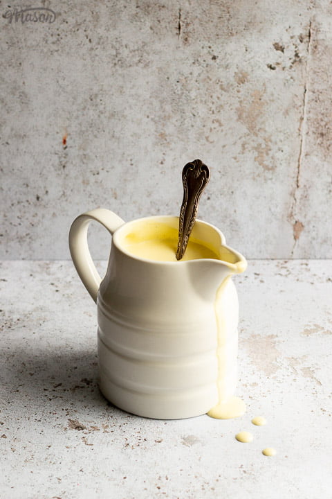 Homemade custard in a white jug with drips running down the front and a silver spoon inside