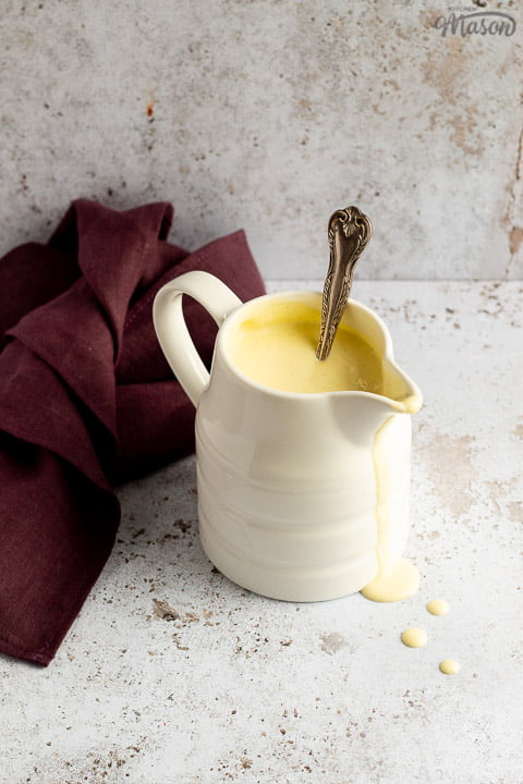 Homemade custard in a white jug on a burgundy linen napkin with drips running down the front and a silver spoon inside