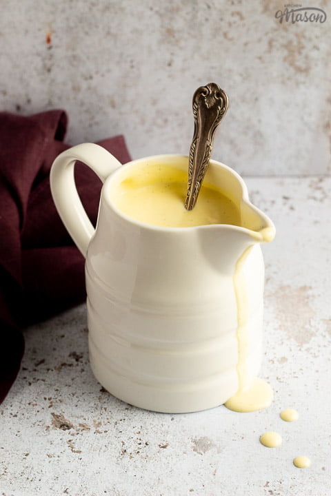 Homemade custard in a white jug on a burgundy linen napkin with drips running down the front and a silver spoon inside