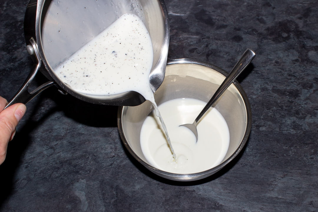 Hot vanilla infused milk being poured into a metal bowl containing a milk and cornflour paste and a metal spoon