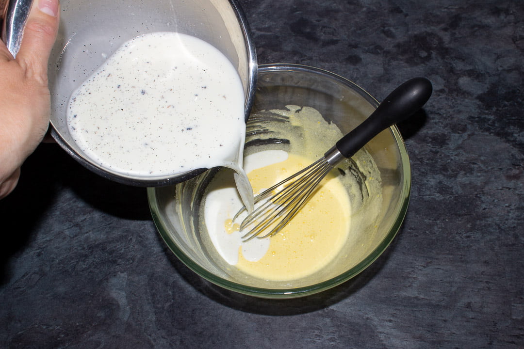 A vanilla infused milk and cornflour mixture being poured into a glass bowl containing an egg yolk and sugar mixture and a whisk
