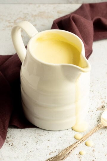 Homemade custard in a white jug on a burgundy linen napkin with drips running down the front and a silver spoon in the background