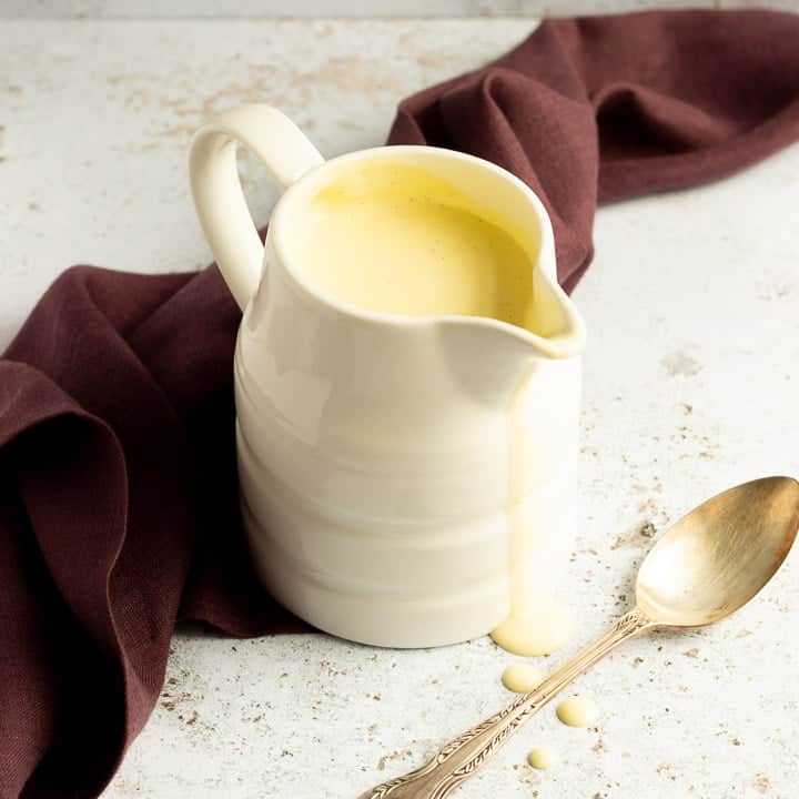 Homemade custard in a white jug on a burgundy linen napkin with drips running down the front and a silver spoon in the background