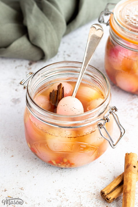 A jar of pickled onions with a fork inside on a white mottled background with a green linen napkin, cinnamon sticks and another jar