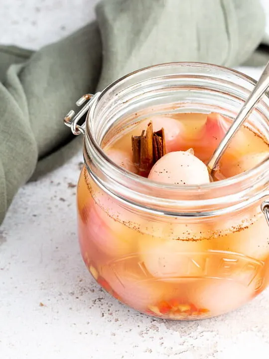 A jar of pickled onions with a fork inside on a white mottled background with a green linen napkin and cinnamon sticks