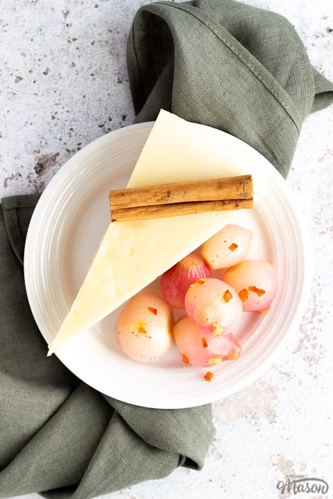 Pickled onions on a white plate with a wedge of pecorino cheese and a cinnamon stick laid on a white mottled background