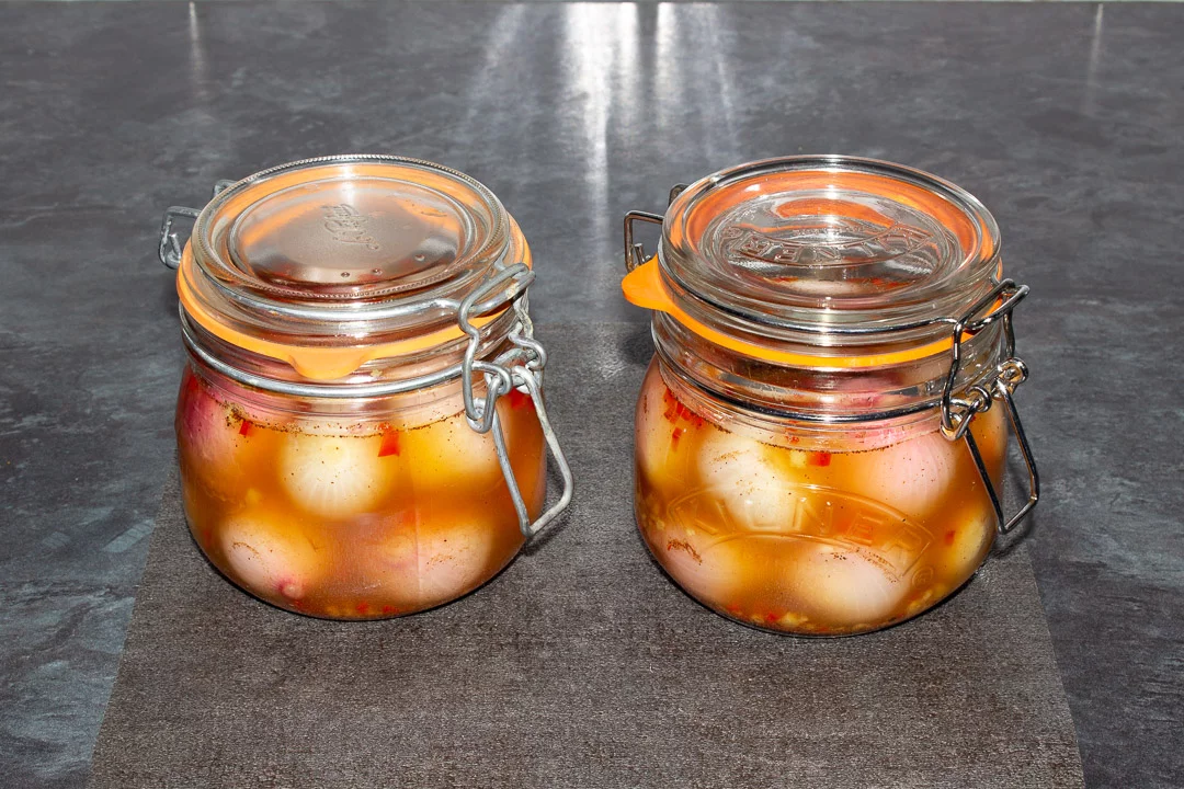 Two sealed jars filled with onions and a spiced pickling liquid on a heat proof mat