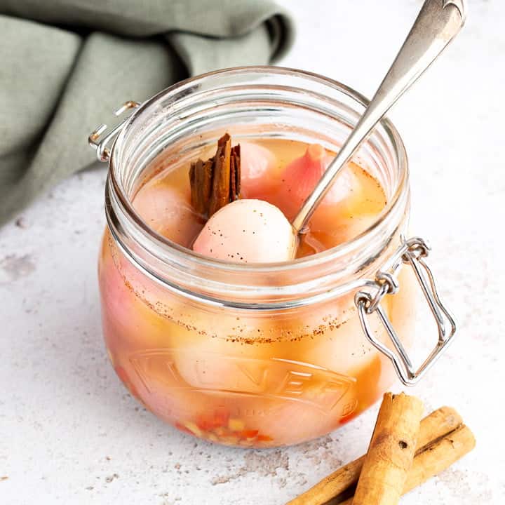 A jar of pickled onions with a fork inside on a white mottled background with a green linen napkin and cinnamon sticks