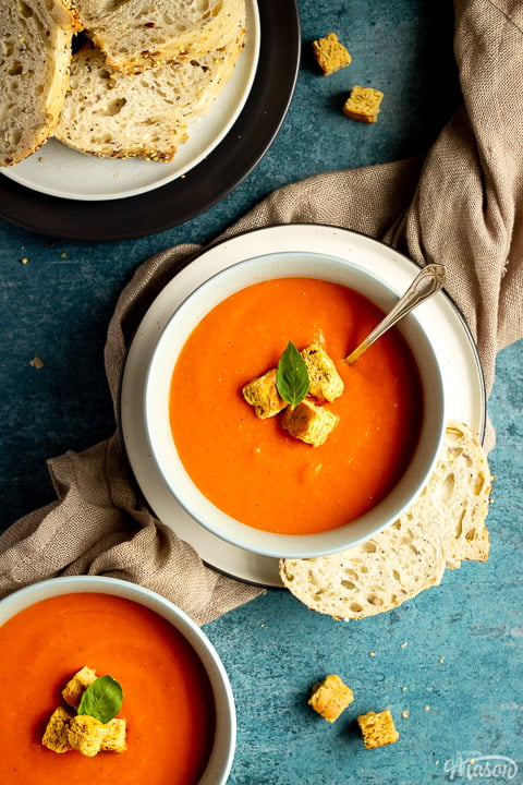 A bowl of tomato soup with croutons, a leaf of basil and a spoon inside, set on a plate on a light brown napkin with a slice of sourdough bread on the side. There is another bowl of soup, a plate with slices of bread and some croutons in the background.