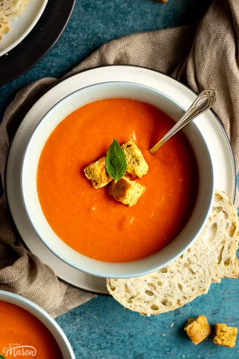 A bowl of tomato soup with croutons, a leaf of basil and a spoon inside, set on a plate on a light brown napkin with a slice of sourdough bread on the side. There is another bowl of soup, a plate with slices of bread and some croutons in the background.