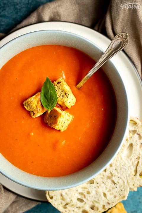 A bowl of tomato soup with croutons, a leaf of basil and a spoon inside, set on a plate on a light brown napkin with a slice of sourdough bread on the side. There are also some croutons in the background.