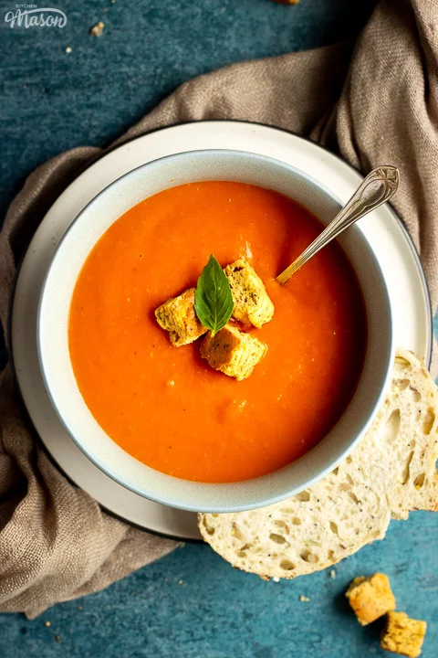 A bowl of tomato soup with croutons, a leaf of basil and a spoon inside, set on a plate on a light brown napkin with a slice of sourdough bread on the side. There are also some croutons in the background.