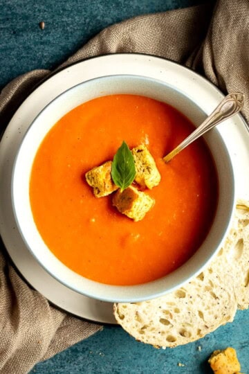 A bowl of tomato soup with croutons, a leaf of basil and a spoon inside, set on a plate on a light brown napkin with a slice of sourdough bread on the side. There are also some croutons in the background.