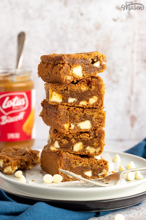 5 White chocolate Biscoff blondies in a stack on top of two plates with a fork on the side. An open jar of Biscoff spread with a spoon in it sits in the background and a blue linen napkin.