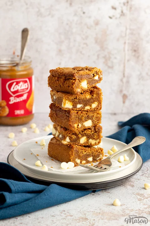 5 White chocolate Biscoff blondies in a stack on top of two plates with a fork on the side. An open jar of Biscoff spread with a spoon in it sits in the background and a blue linen napkin.
