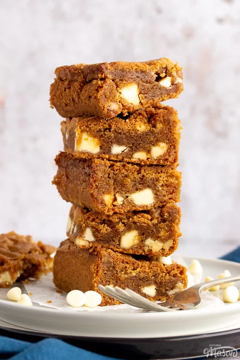 5 White chocolate Biscoff blondies in a stack on top of two plates with a fork on the side. A blue linen napkin sits underneath.