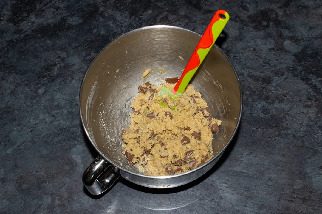 Chocolate chip cookie dough in the bowl of en electric stand mixer with a green rubber spatula