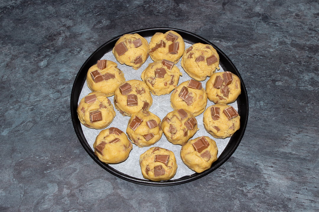 Balls of chocolate chip cookie dough (with extra chocolate chips on top) on a black plate lined with baking paper