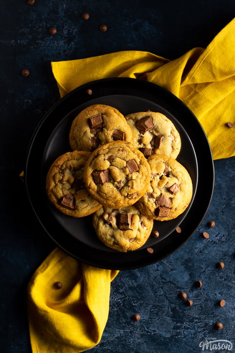 6 Chewy chocolate chip cookies on 2 stacked black plates set over a yellow linen napkin. With a deep blue marbled backdrop and chocolate chips scattered around.