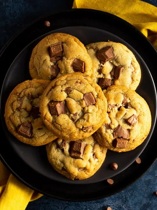 6 Chewy chocolate chip cookies on 2 stacked black plates set over a yellow linen napkin. With a deep blue marbled backdrop and chocolate chips scattered around.