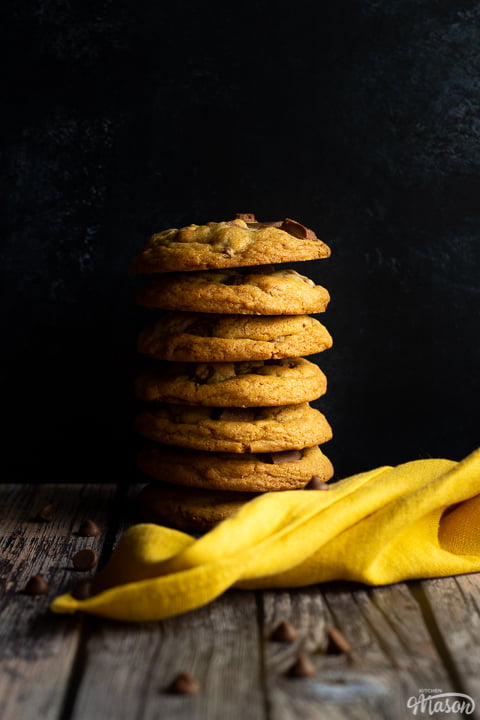 A stack of 7 chewy chocolate chip cookies against a deep blue marbled backdrop. There is a yellow linen napkin and chocolate chips scattered around it.