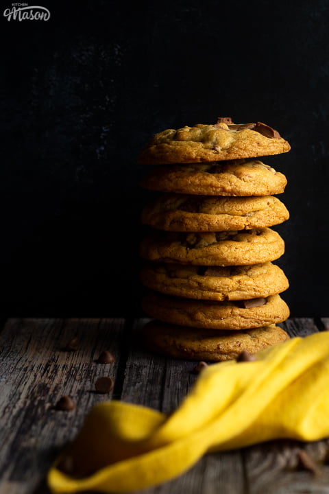 A stack of 7 chewy chocolate chip cookies against a deep blue marbled backdrop. There is a yellow linen napkin and chocolate chips scattered around it.