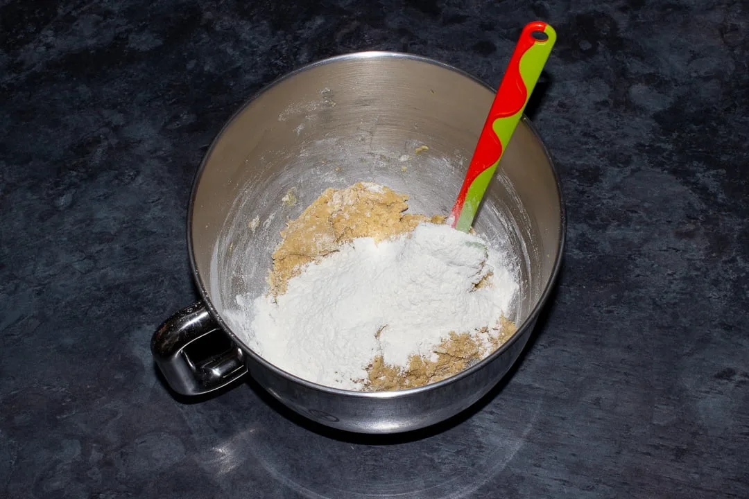 Cookie dough with the remaining flour mixture on top in the bowl of an electric stand mixer with a green rubber spatula