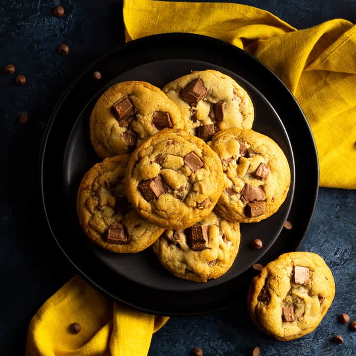 6 Chewy chocolate chip cookies on 2 stacked black plates set over a yellow linen napkin. With a deep blue marbled backdrop with more cookies and chocolate chips scattered around.