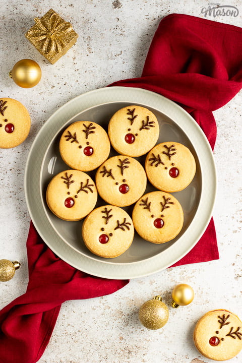 A plate of Reindeer cookies set on a second plate and a red linen napkin. Set on an off white backdrop with golden baubles, golden presents and more reindeer cookies in the background.