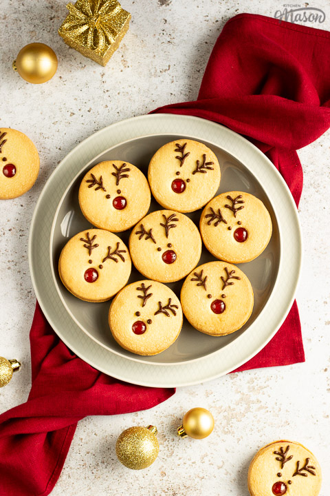 A plate of Reindeer cookies set on a second plate and a red linen napkin. Set on an off white backdrop with golden baubles, golden presents and more reindeer cookies in the background.
